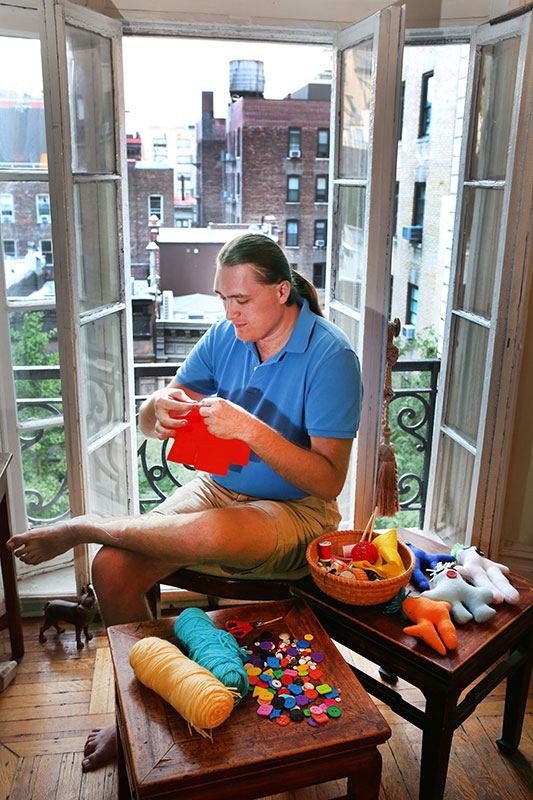 Sam Montgomery hand-sewing a doll beside the window in his New York City home